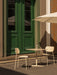 A sunlit patio with an ivory Nook Dining Table, two Co Dining Chairs, and a Column Portable lamp before a large green door.