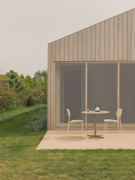 Outdoor patio with a Nook Dining Table, two Co Dining Chairs, and a Carrie Lamp, set against a modern wood-panelled building.