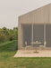 Outdoor patio with a Nook Dining Table, two Co Dining Chairs, and a Carrie Lamp, set against a modern wood-panelled building.