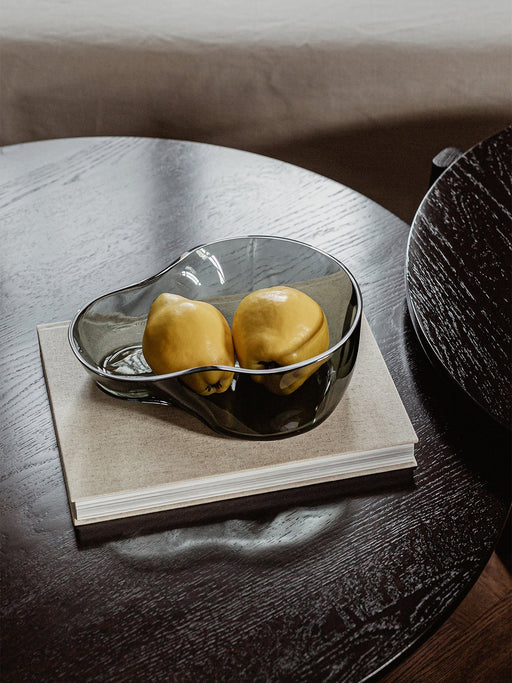 A close-up of the Aer Bowl in smoked glass, holding two yellow quinces. The bowl rests on a book on top of a dark wood table.