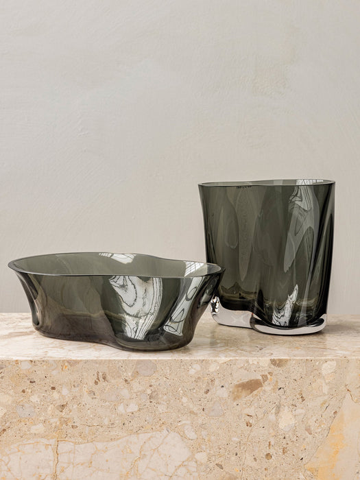 The Aer Vase and Aer Bowl in smoky grey glass, both with an organic, rippled shape, sit on a beige marble plinth.