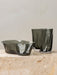 The Aer Vase and Aer Bowl in smoky grey glass, both with an organic, rippled shape, sit on a beige marble plinth.