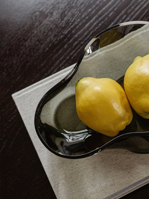 A close-up of the organically shaped Aer Bowl in smoked glass, holding two yellow lemons on a grey book over a dark table.