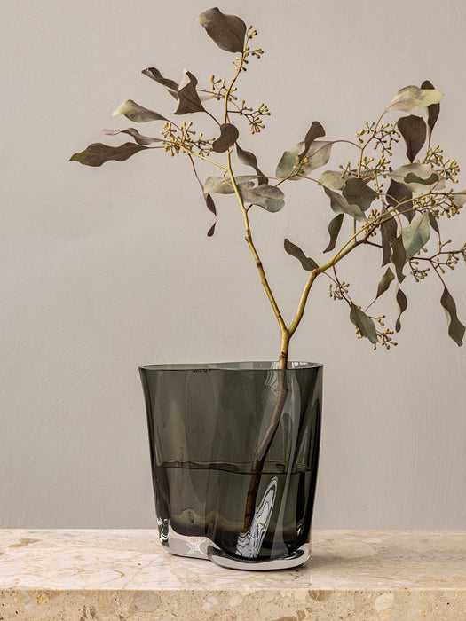 A close-up of the smokey grey glass Aer Vase with its rippled silhouette, holding a eucalyptus branch on a marble surface.