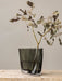 A close-up of the smokey grey glass Aer Vase with its rippled silhouette, holding a eucalyptus branch on a marble surface.