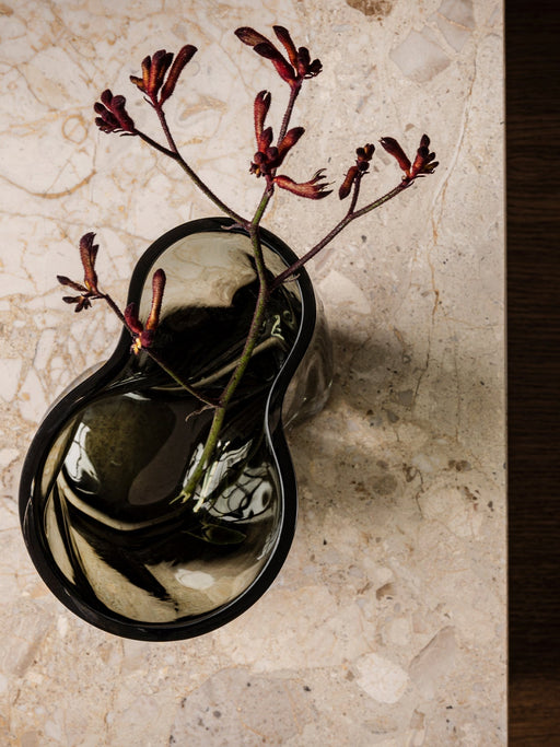A top-down view of the dark, rippled glass Aer Vase holding a stem of fuzzy, deep red Kangaroo Paws on a marble surface.