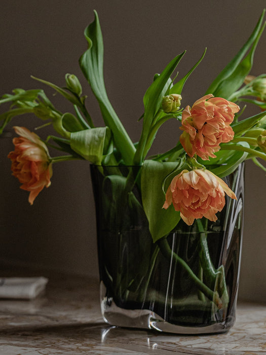 A close-up of the dark smoked glass Aer Vase, filled with a bouquet of orange ruffled tulips, sitting on a marble surface.