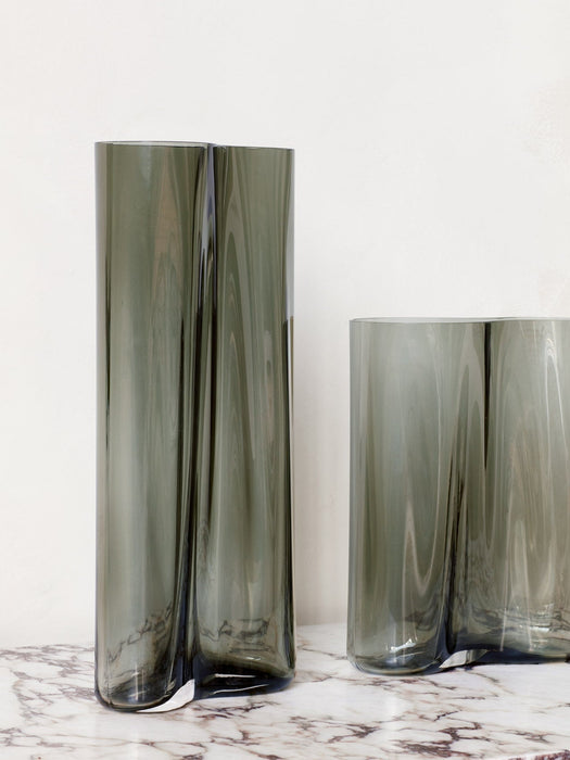 A tall and a short Aer Vase in undulating, smoky grey glass, displayed on a white marble tabletop against a plain wall.