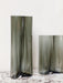 A tall and a short Aer Vase in undulating, smoky grey glass, displayed on a white marble tabletop against a plain wall.
