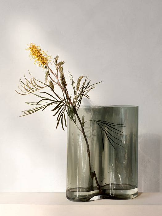 A tall, smoked grey glass Aer Vase with a wavy, organic shape, holding a single stem with a yellow bottlebrush flower.