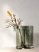 A tall, smoked grey glass Aer Vase with a wavy, organic shape, holding a single stem with a yellow bottlebrush flower.