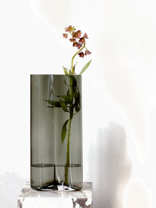 A tall Aer Vase in smoked grey glass holds a single stem of small, purple flowers. The vase is positioned on a marble plinth.