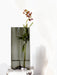 A tall Aer Vase in smoked grey glass holds a single stem of small, purple flowers. The vase is positioned on a marble plinth.