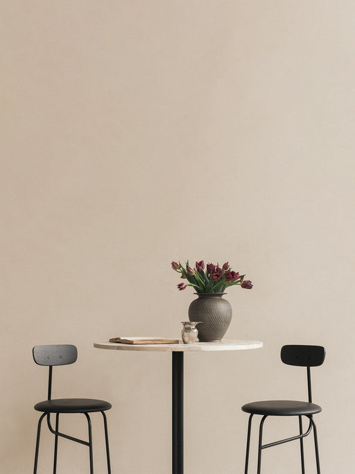 Two black Afteroom Bar Chairs flank a marble-topped Harbour Column Bar Table holding a vase of flowers against a beige wall.
