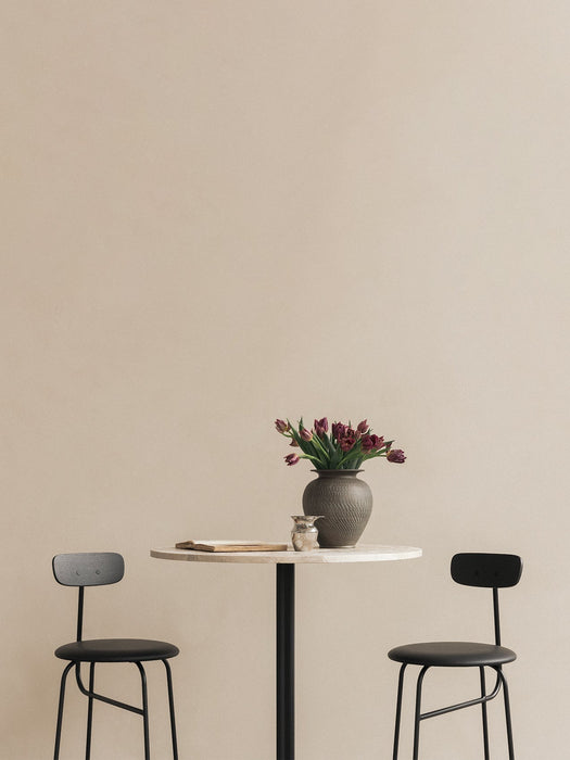 Two black Afteroom Bar Chairs flank a marble-topped Harbour Column Bar Table holding a vase of flowers against a beige wall.