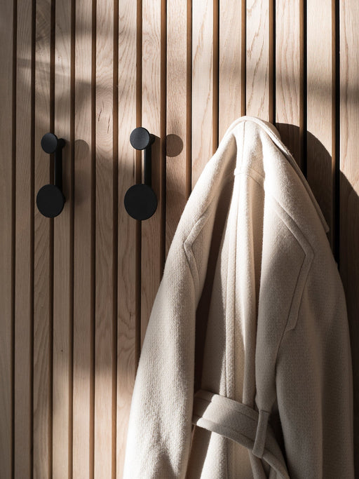 Two black minimalist Knobs wall hooks mounted on a light oak slatted wall, with a cream wool coat hanging from one hook.