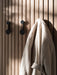 Two black minimalist Knobs wall hooks mounted on a light oak slatted wall, with a cream wool coat hanging from one hook.