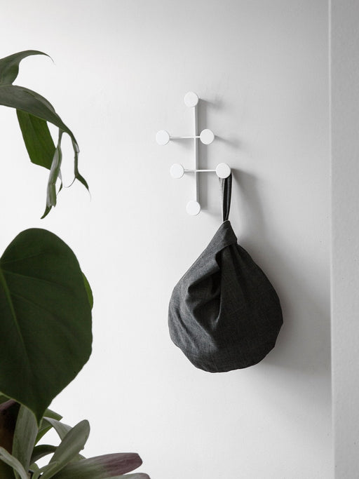 A white Afteroom Coat Hanger mounted on a white wall, with a grey knot bag hanging from a peg next to a large green leaf.