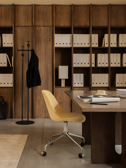 An office with an Androgyne Table, a yellow Harbour Chair, Afteroom Coat Stand, and Nonna Lamp against dark wood shelving.