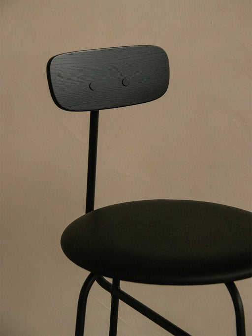 A close-up of the black Afteroom Counter Chair, Veneer, against a beige wall, focusing on the grained wood backrest.