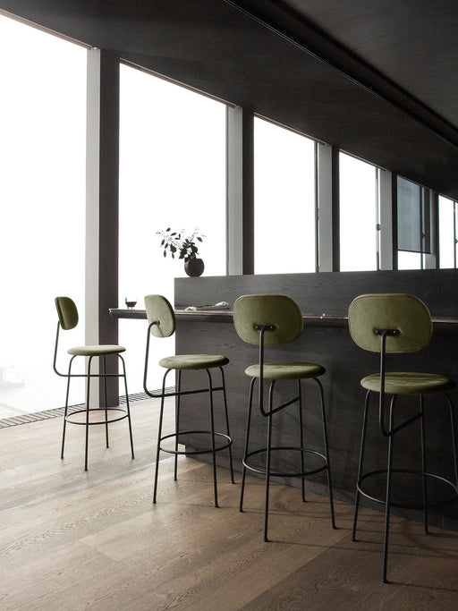 Four green upholstered Afteroom Bar Chair Plus at a dark bar in a modern room with large windows and light wood floors.