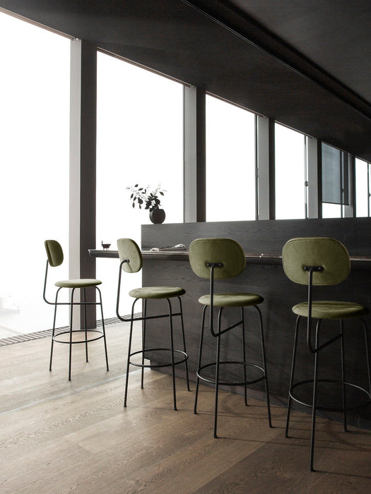 Four green upholstered Afteroom Bar Chair Plus at a dark bar in a modern room with large windows and light wood floors.