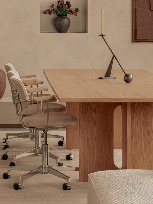 A light oak Androgyne Dining Table is shown from the side with two upholstered office chairs and a minimalist candlestick.