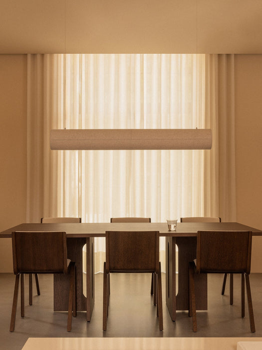 A long dark wood Androgyne Dining Table with matching Ready Dining Chairs, illuminated by a Hashira Linear Pendant Lamp.