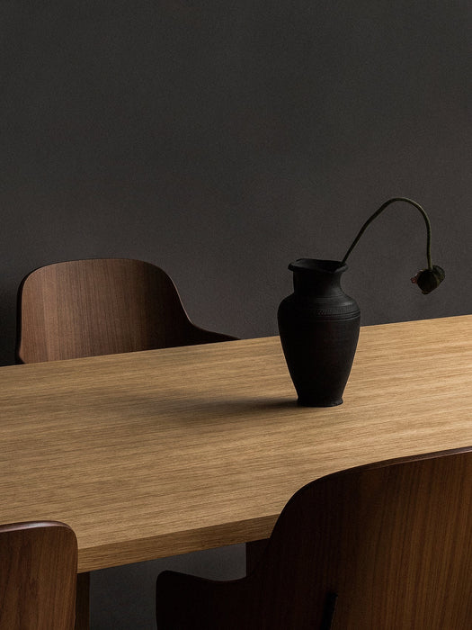 A corner of the light oak Androgyne Dining Table with a matte black vase holding a single wilting flower, against dark wall.