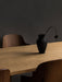 A corner of the light oak Androgyne Dining Table with a matte black vase holding a single wilting flower, against dark wall.