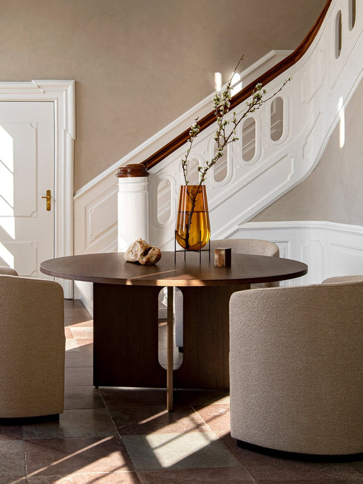 A round, dark oak Androgyne Dining Table is styled with an amber vase and surrounded by chairs in a grand, sunlit entryway.