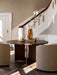 A round, dark oak Androgyne Dining Table is styled with an amber vase and surrounded by chairs in a grand, sunlit entryway.