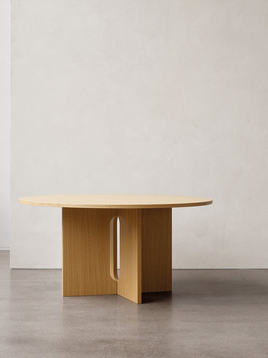 A light oak Androgyne Dining Table with a round top and a cross-shaped base with a vertical slot, against a white wall.
