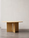 A light oak Androgyne Dining Table with a round top and a cross-shaped base with a vertical slot, against a white wall.