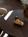A close-up of the round, dark-stained oak Androgyne Dining Table, styled with a vase of blossoms, a stone, and a brass cube.