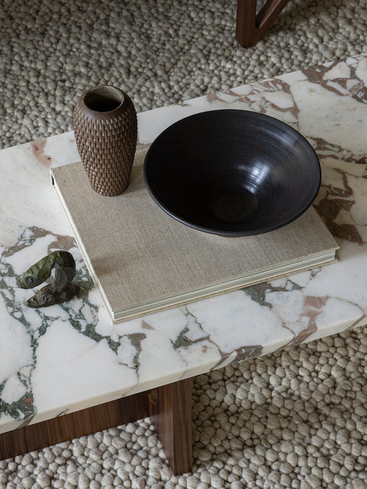 Close-up of the Androgyne Lounge Table with a marble top and wood base, styled with a book, vase, and bowl on a textured rug.