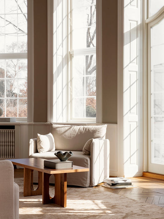 A sunlit living room featuring the wooden Androgyne Lounge Table in front of a beige armchair, with light from large windows.