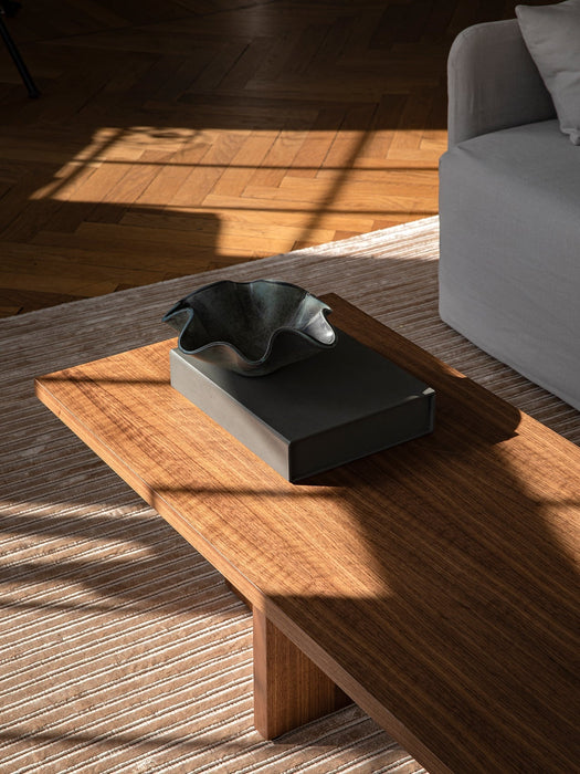 A dark wood Androgyne Lounge Table sits on a striped rug, styled with a book and decorative bowl in a sunlit room.