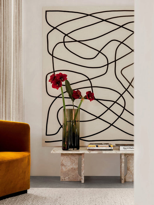 Androgyne Lounge Table in stone, styled with red flowers in a vase, beside a yellow armchair and in front of abstract art.