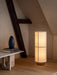 The stone Androgyne Lounge Table with books and a vase, next to an illuminated floor lamp in a room with wooden beams.
