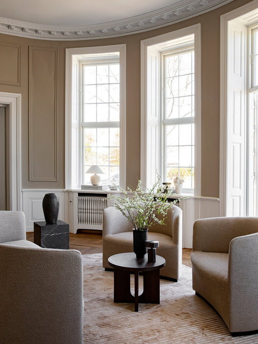 A dark wood Androgyne Side Table in a classic living room with large windows, surrounded by beige chairs on a patterned rug.