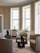 A dark wood Androgyne Side Table in a classic living room with large windows, surrounded by beige chairs on a patterned rug.