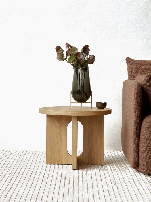 A natural oak Androgyne Side Table styled with a vase of flowers and a small bowl, set beside a brown sofa on a ribbed rug.