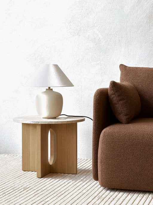 Androgyne Side Table with an oak base and stone top, holding a cream lamp. It sits next to a brown sofa on a striped rug.