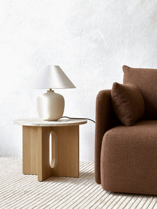 Androgyne Side Table with an oak base and stone top, holding a cream lamp. It sits next to a brown sofa on a striped rug.