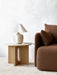 Androgyne Side Table with an oak base and stone top, holding a cream lamp. It sits next to a brown sofa on a striped rug.