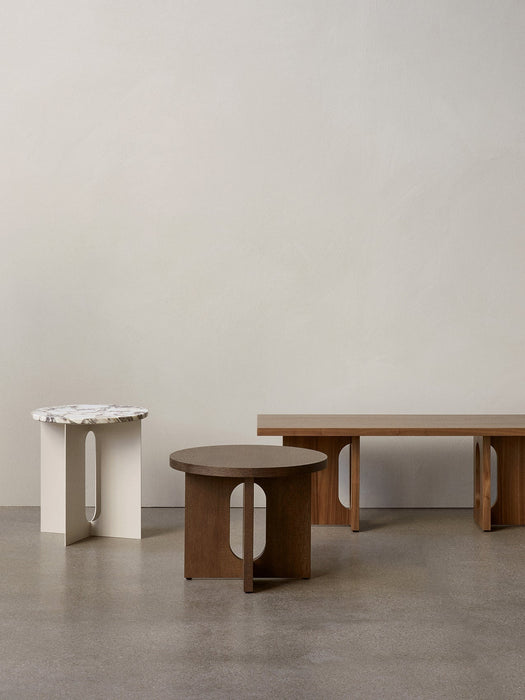 Three Androgyne tables on a grey floor: a white base/marble top side table, a dark oak side table, and a light oak bench.