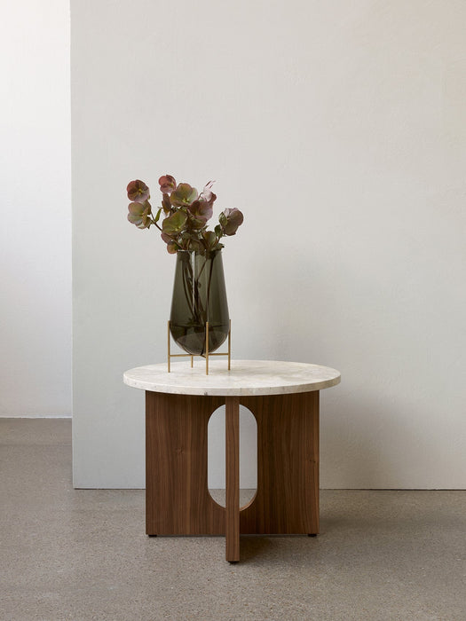 The Androgyne Side Table with a walnut base and Kunis Breccia stone top, styled with a dark glass vase and flowers.