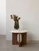 The Androgyne Side Table with a walnut base and Kunis Breccia stone top, styled with a dark glass vase and flowers.
