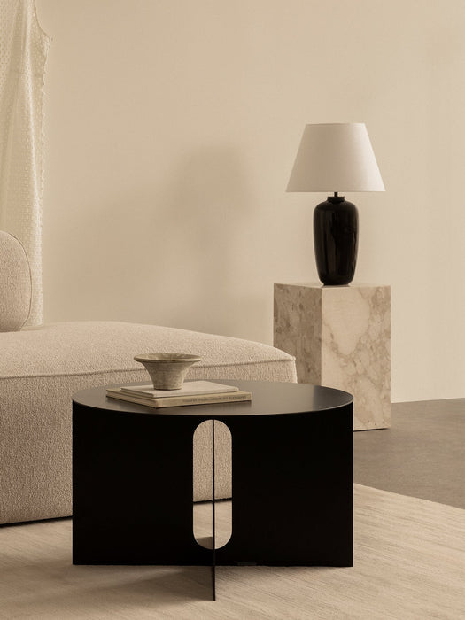 A black Androgyne Side Table beside an Eave Modular Sofa. In the background, a Torso Table Lamp rests on a marble Plinth.